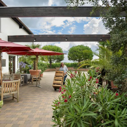 Ein modernes Haus, mit gepflasterter Terasse und zwei Tischen, mit roten Sonnenschirmen. Davor läuft eine Frau und schieb ein Weinfass vor sich her.