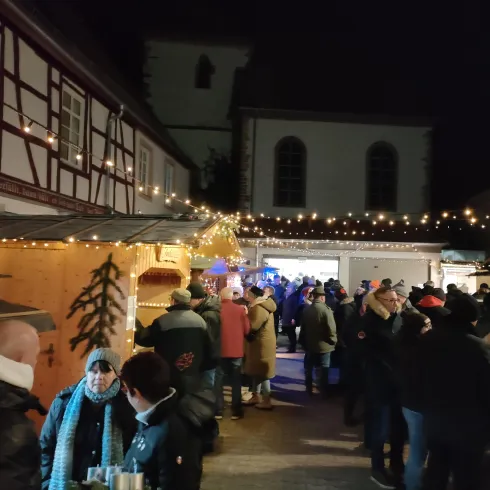 Ein festlicher Marktplatz mit stimmungsvoller Beleuchtung und vielen Menschen. Stände bieten verschiedene Waren an und die Atmosphäre ist gemütlich.