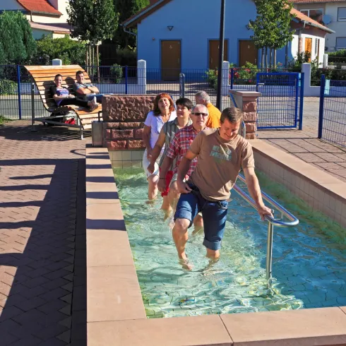 Eine Gruppe von Menschen geht durch ein Tretbecken in einer sonnigen Umgebung. Im Hintergrund entspannen einige Personen auf Bänken.