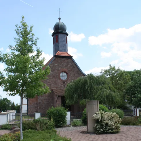 Eine kleine Kirche umgeben von Grünanlagen und Bäumen. Der Weg zum Gebäude ist mit Steinen gepflastert.