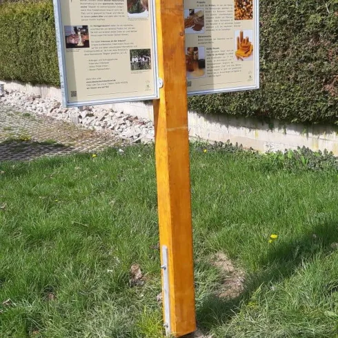 Ein Informationsschild steht auf einer Wiese, umgeben von Gras und Steinen. Im Hintergrund sind Bäume und ein Gebäude zu sehen.