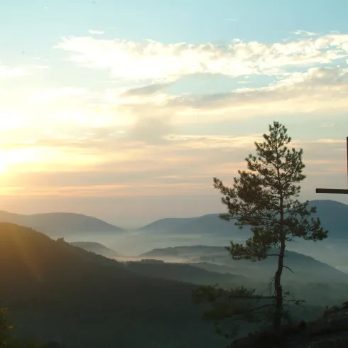 Eine ruhige Landschaft mit sanften Hügeln und einem Kreuz im Vordergrund. Der Sonnenaufgang taucht die Szene in warmes Licht.
