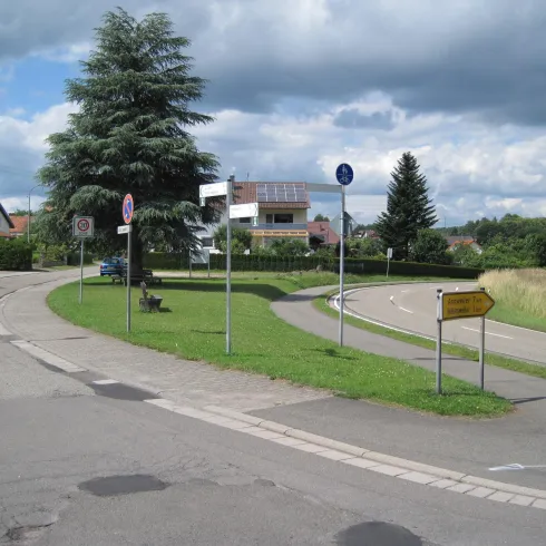 Eine ruhige Straße mit einem Bogen und Grünflächen. Im Hintergrund sind einige Häuser und ein großer Baum zu sehen.