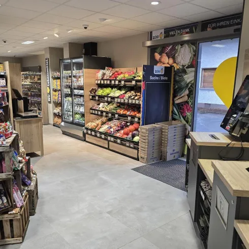 Ein moderner Verkaufsraum mit verschiedenen Lebensmittelregalen und Snacks. Auf der rechten Seite sind Obst und Gemüse, während sich auf der linken Seite einige Snacks in Holzkisten befinden.