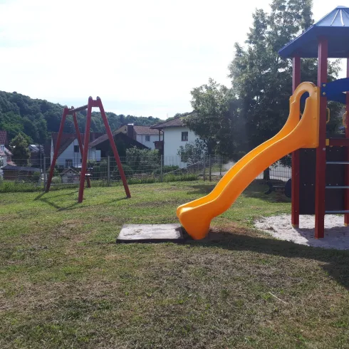 Ein Spielplatz mit einer Rutsche in orange und einer Wippe. Im Hintergrund sind grüne Bäume und einige Häuser sichtbar.