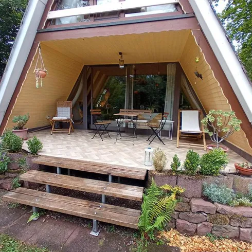 Ein gemütliches Ferienhaus mit einem modernen A-Rahmen und einer einladenden Terrasse. Die Umgebung ist grün und schön gestaltet mit Pflanzen und Sitzgelegenheiten.
