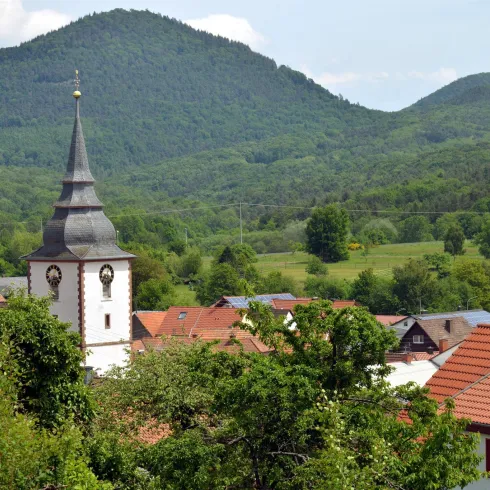 Eine malerische Landschaft mit einem kleinen Dorf und einer Kirche im Vordergrund. Im Hintergrund erheben sich grüne Hügel unter einem klaren Himmel.