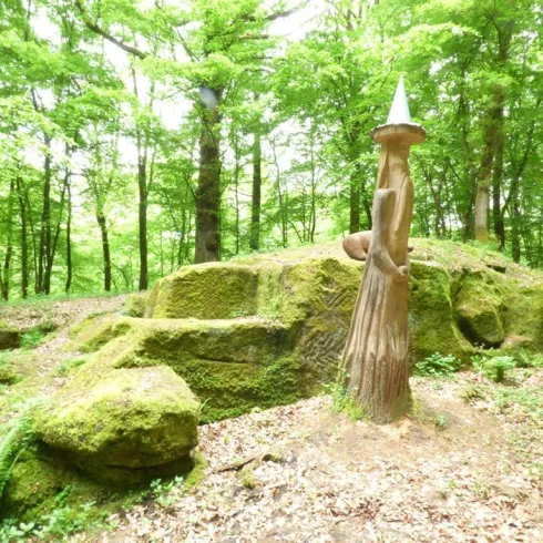 Eine Holzskulptur in einem grünen Wald steht auf einem moosbedeckten Felsen. Die Umgebung ist lebendig und voller Bäume.