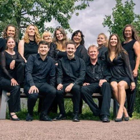 Eine Gruppe von 14 Personen in eleganter schwarzer Kleidung sitzt und steht auf einer Bank im Freien. Die Umgebung ist grün mit Bäumen im Hintergrund.