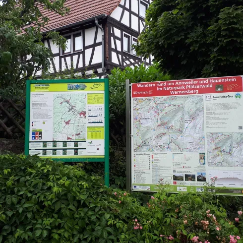 Zwei Informationstafeln über Wanderwege stehen vor einem traditionellen Fachwerkhaus. Die Umgebung ist grün und blühend.