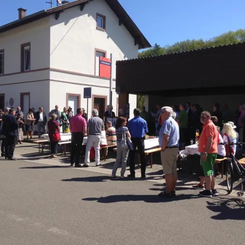 Eine Gruppe von Menschen versammelt sich vor einem Gebäude an einem sonnigen Tag. Tische sind aufgestellt, und es scheint eine Veranstaltung oder ein Treffen zu sein.
