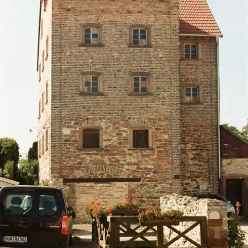 Ein altes, zweigeschossiges Gebäude aus Backstein mit einem roten Ziegeldach. Vor dem Haus stehen Blumen und ein geparkter schwarzer Wagen.