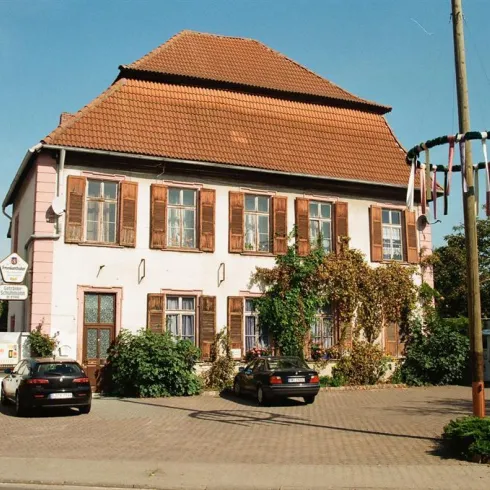 Ein charmantes Gebäude mit einem roten Ziegeldach und großen Fenstern. Vor dem Haus stehen einige Autos und es gibt Pflanzen, die die Fassade schmücken.