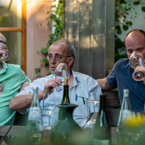 Drei Männer sitzen an einem Tisch und genießen Wein. Im Hintergrund sind grüne Pflanzen und eine entspannte Atmosphäre zu sehen.