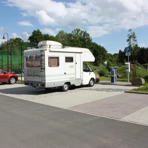 Ein Wohnmobil steht auf dem Parkplatz neben einem roten Auto. Im Hintergrund sind Bäume und ein Tennisplatz zu sehen.