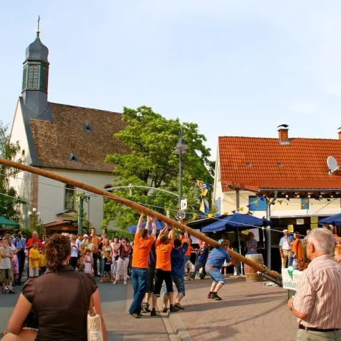 Eine Menschenmenge feiert auf einer Straße mit einem traditionellen Fest. Einige Teilnehmer heben einen langen Baumstamm, während im Hintergrund ein Gebäude mit einem Kirchturm zu sehen ist.