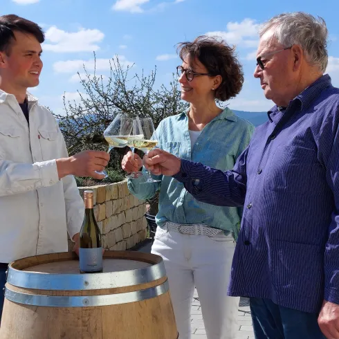 Drei Personen halten Gläser und stoßen an. Im Hintergrund sind Weinberge und ein blauer Himmel zu sehen.