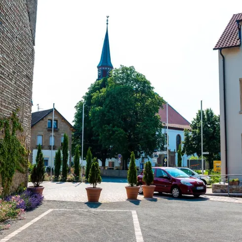 Eine ruhige Straße mit blühenden Pflanzen und Töpfen. Im Hintergrund ist ein Kirchturm sichtbar, umgeben von Gebäuden.