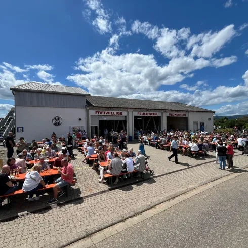 Eine große Menschenmenge sitzt an den Tischen vor einem Gebäude. Das Wetter ist sonnig mit vielen Wolken am Himmel.