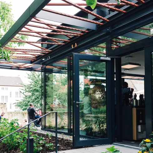 Ein modernes Gebäude mit großen Glasfenstern und einem darüber liegenden Dach. Draußen sind Menschen und Pflanzen zu sehen, was eine einladende Atmosphäre schafft.