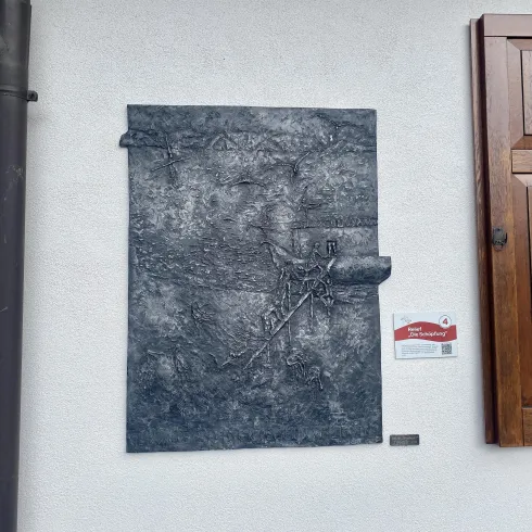 Eine Metallplatte mit einer strukturierten Oberfläche hängt an einer weißen Wand. Neben der Platte befindet sich ein Holzfenster und ein Schild mit Informationen.