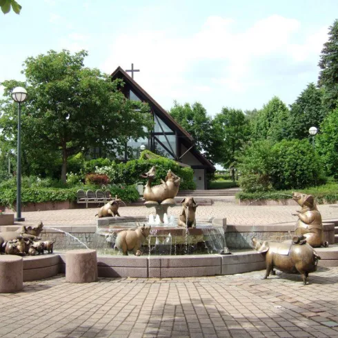 Ein Brunnen mit bronzenen Schweinfiguren in einem begrünten Platz. Im Hintergrund ist ein modernes Gebäude mit großen Fenstern zu sehen.