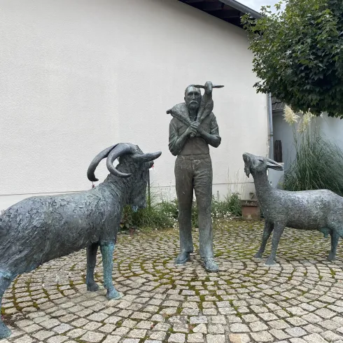 Eine Bronzeskulptur eines Mannes mit einer Axt, umgeben von zwei Ziegen. Die Szene ist von einem gepflasterten Platz umgeben.