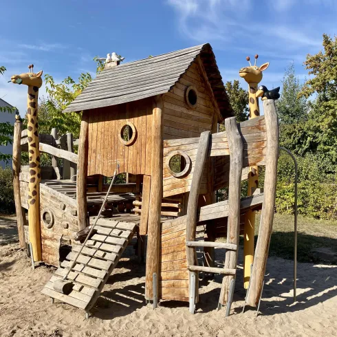Ein kreatives Spielhaus aus Holz mit einer Rutsche und Kletterelementen. Es hat zwei kleine Giraffenfiguren an den Seiten und ist von Bäumen umgeben.