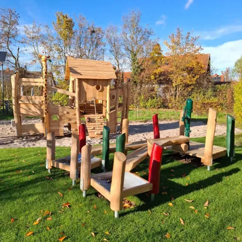 Ein exotischer Spielplatz aus Holz mit verschiedenen Kletterelementen. Umgeben von grünem Rasen und bunten Bäumen im Hintergrund.