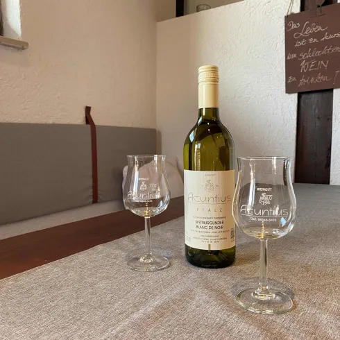 Eine Flasche Weißwein steht auf einem Tisch mit zwei Weingläsern. Im Hintergrund ist eine Wand mit einem handschriftlichen Schild zu sehen.