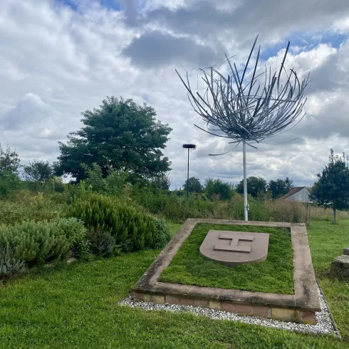 Eine künstlerische Skulptur in einem grünen Park mit einem großen, geschwungenen Drahtaufbau. Im Hintergrund sind Bäume und ein bewölkter Himmel zu sehen.