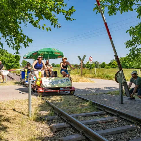Eine Gruppe von Personen fährt mit einer Fahrraddraisine entlang der Schienen. Die Szene ist von viel Grün und sonnigem Wetter umgeben.