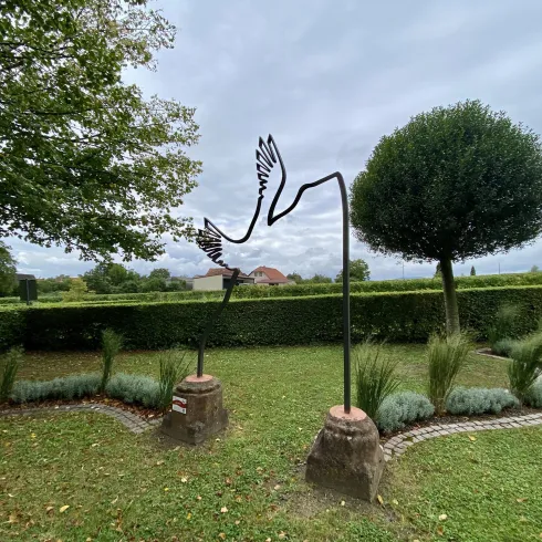 Eine moderne Skulptur von fliegenden Vögeln steht in einem gepflegten Garten. Im Hintergrund sind Bäume und eine grüne Hecke zu sehen.