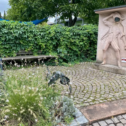 Eine kleine Parkanlage mit einer steinernen Statue und einer Bank. Umgeben von grünen Sträuchern und blühenden Pflanzen.