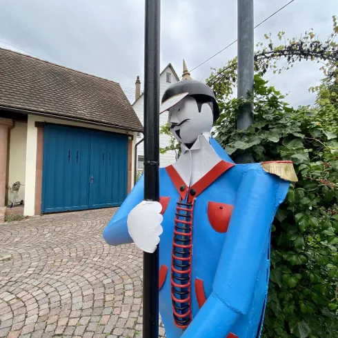 Ein bunter Straßenfigurenmann steht neben einem Laternenpfahl. Im Hintergrund sind ein blaues Tor und ein grünes Gebüsch zu sehen.