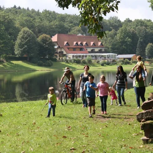Eine Gruppe von Menschen spaziert am Ufer eines Sees. Im Hintergrund ist ein nettes Gebäude und Bäume zu sehen.