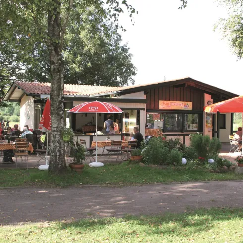 Ein einladendes Café mit Terrassensitzplätzen und roten Sonnenschirmen. Umgeben von Bäumen und einer grünen Wiese.