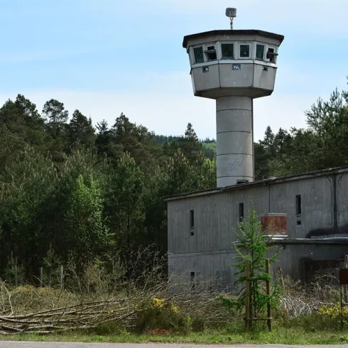 Ein verlassener Gebäudekomplex mit einem Wachturm und umgebenden Bäumen. Die Natur hat den Ort zurückerobert, mit wilden Pflanzen und verwildertem Gelände.