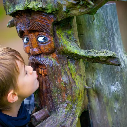 Ein Junge küsst eine Holzskulptur eines Gesichts, das an einem Baumstamm geschnitzt ist. Die Skulptur hat leuchtende Augen und ein freundliches Lächeln.