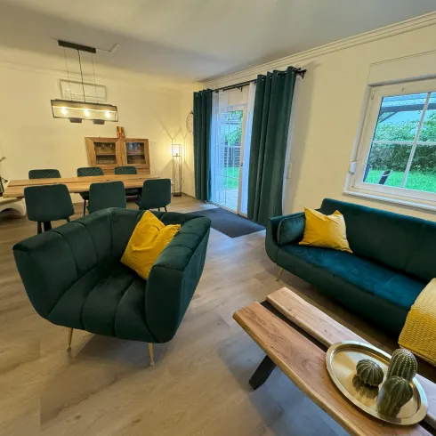 Ein modernes Wohnzimmer mit bequemen Möbeln in grünen und gelben Farbtönen. Große Fenster sorgen für viel Tageslicht und Ausblick auf den Garten.