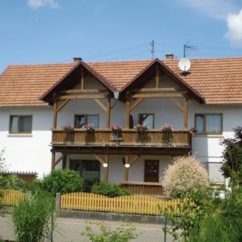 Ein charmantes Haus mit einem Holzbalcony und blühenden Pflanzen. Die Umgebung ist grün und gut gepflegt.