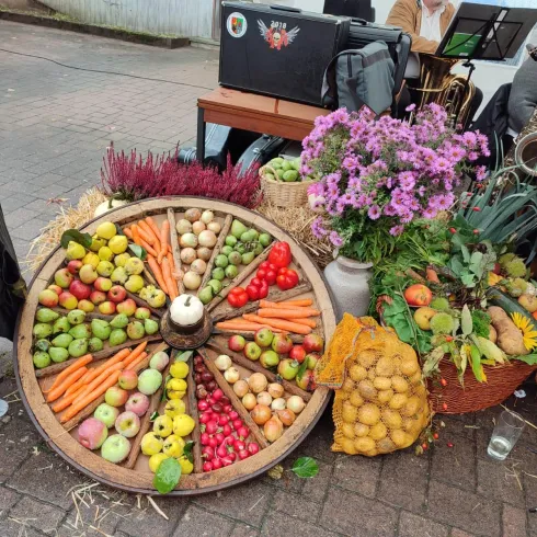 Ein dekorativer Obst- und Gemüsehaufen auf einem Wagenrad, umgeben von bunten Blumen. Die Frucht- und Gemüseanordnung zeigt eine Vielzahl von Farben und Formen.
