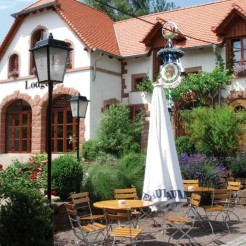 Ein charmantes Gebäude mit roten Ziegeln und Fensterläden. Im Vordergrund befindet sich ein Garten mit Tischen und Stühlen umgeben von bunten Blumen.
