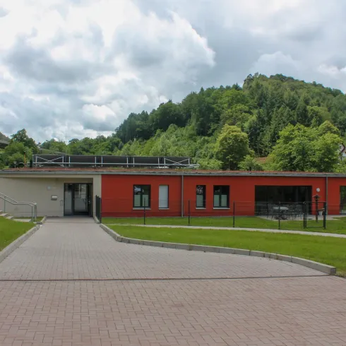 Ein modernes Gebäude in einer ruhigen Umgebung, umgeben von grünen Hügeln. Der Himmel ist bewölkt, und die Einfahrt ist gepflastert.