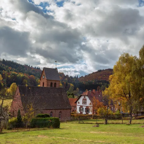 Eine malerische Landschaft mit einer kleinen Kirche und traditionell-bäuerlichen Häusern. Im Hintergrund sind sanfte Hügel und bunten Herbstbäume zu sehen.