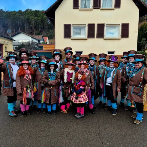 Eine Gruppe von fröhlichen Menschen in bunten Kostümen steht auf einer Straße vor einem Gebäude. Sie tragen Hüte und viele haben bemalte Gesichter.