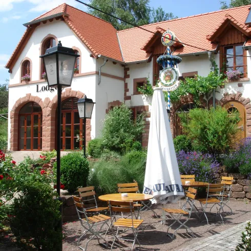 Ein charmantes Haus mit rotem Dach umgeben von bunten Blumen und grünen Pflanzen. Auf der Terrasse stehen Tische mit Stühlen und einem Sonnenschirm.