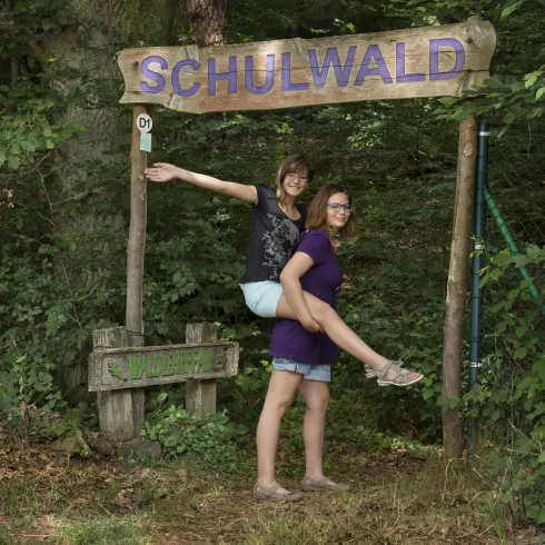 Schild aus Holz mit Aufschrift Schulwald darunter 2 Mädchen die den Schulwald betreten
