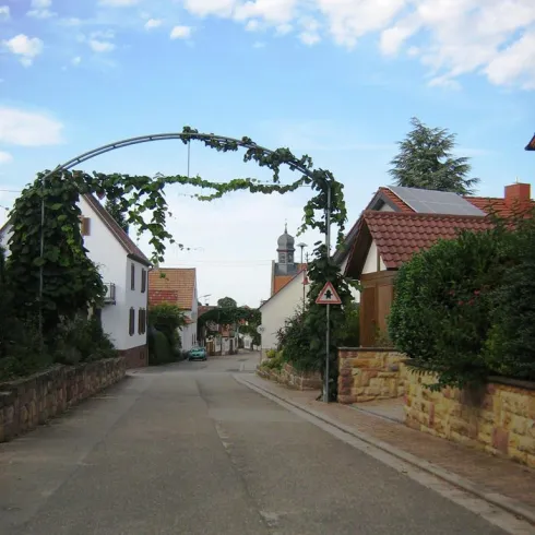 Eine malerische Straße mit einem begrünten Tor und typischen Häusern. Im Hintergrund ist ein Kirchturm zu sehen.