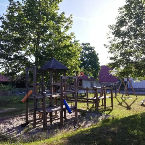 Ein Spielplatz mit Holzstrukturen und Rutschen, umgeben von grünen Bäumen. Der Bereich ist sonnig und bietet viel Platz zum Spielen.